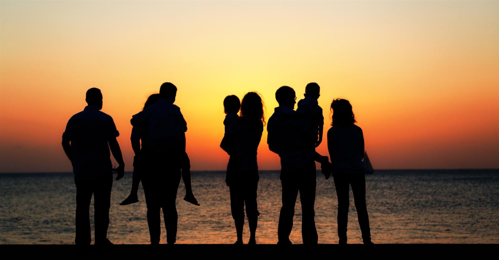 Silhuetas de uma família, com adultos e crianças no colo, vendo o pôr do sol à beira-mar