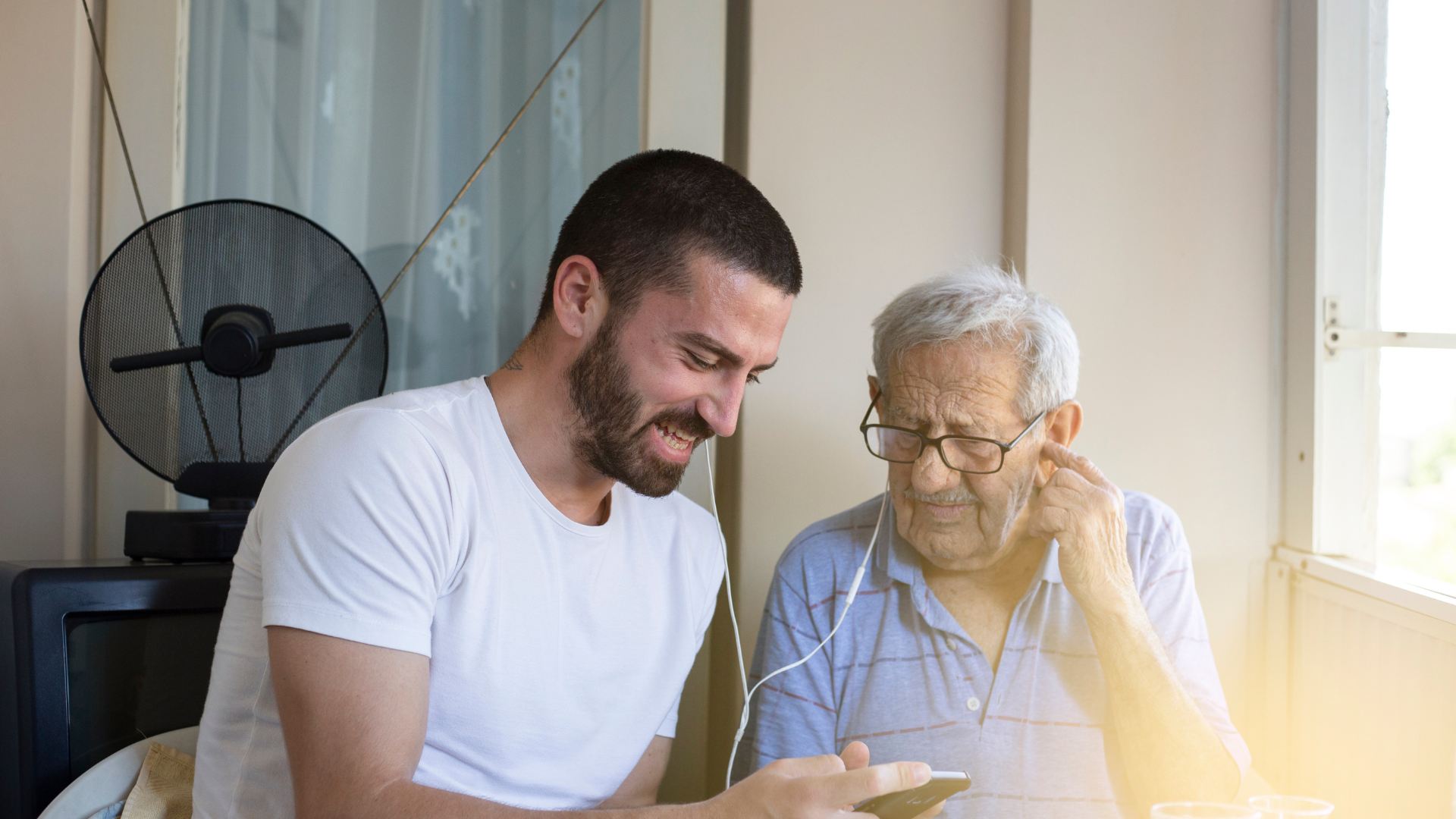Um jovem e um homem de cabelo branco ouvindo juntos uma gravação no celular, com fones de ouvido